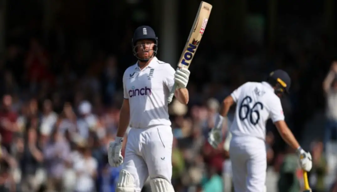 Joe Root celebrating a century with his bat raised to the crowd and teammates applauding from the pavilion.